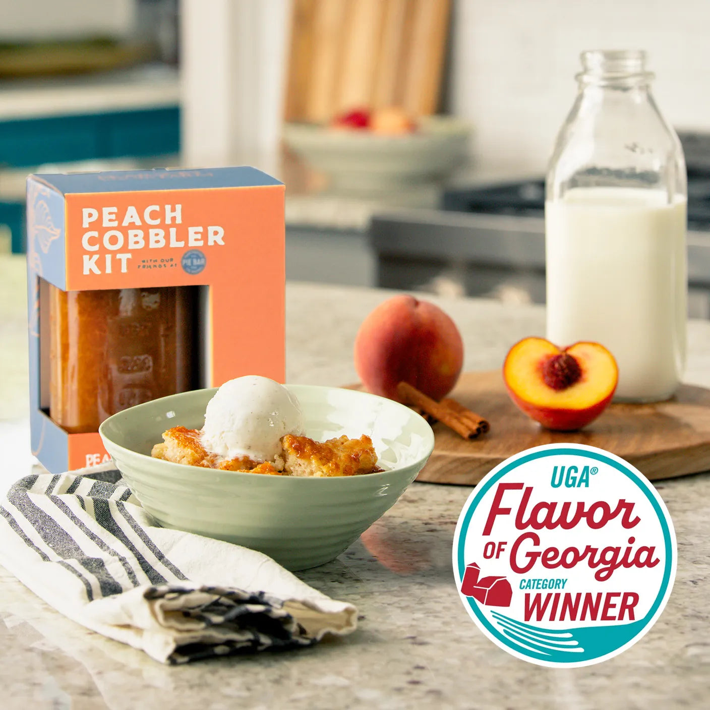 Peach Cobbler Kit box with peach cobbler in bowl topped with ice cream. Sliced peach, cinnamon sticks and bottle of milk on wooden board. Flavor of Georgia winner logo