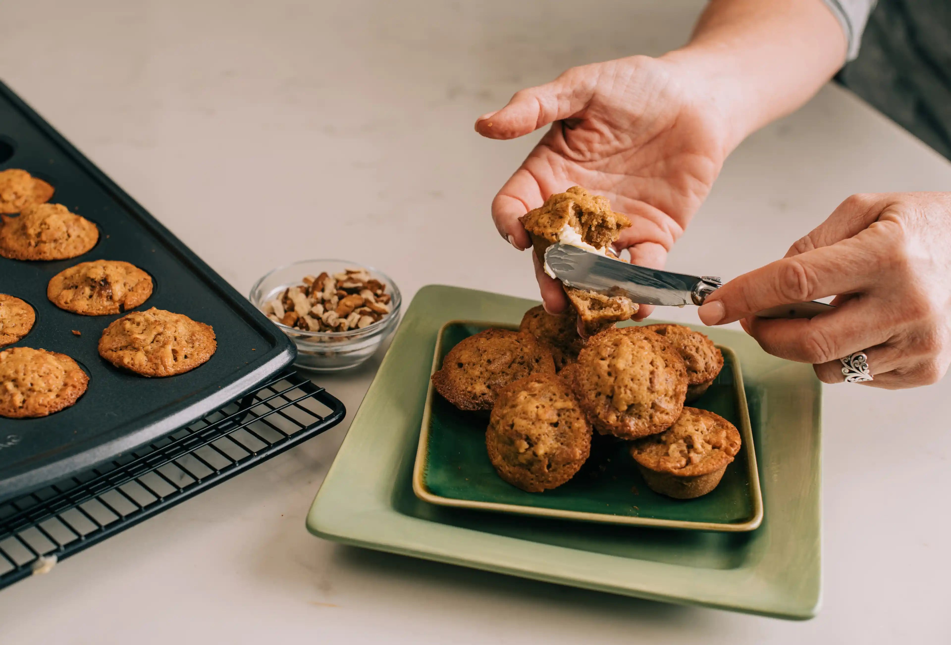 Person serving muffins from a tray onto a green plate with a bowl of nuts on a light-colored surface.