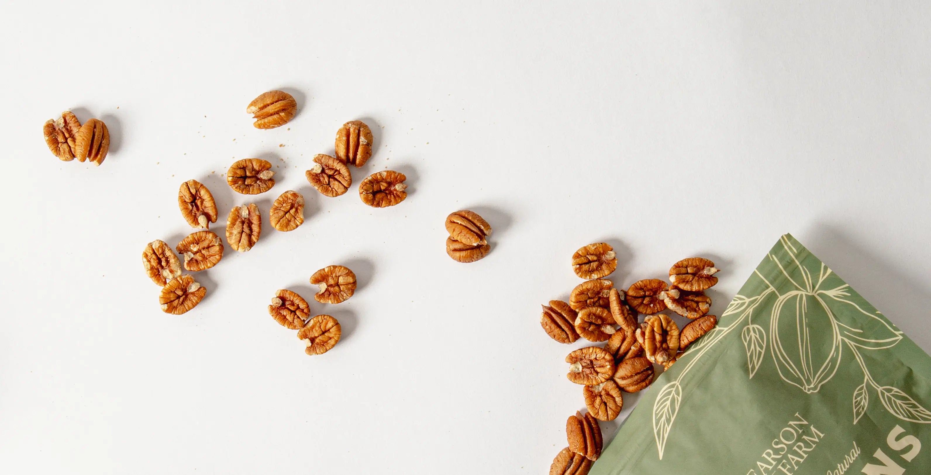 Pecans scattered on a white surface with a green bag featuring pecan design.